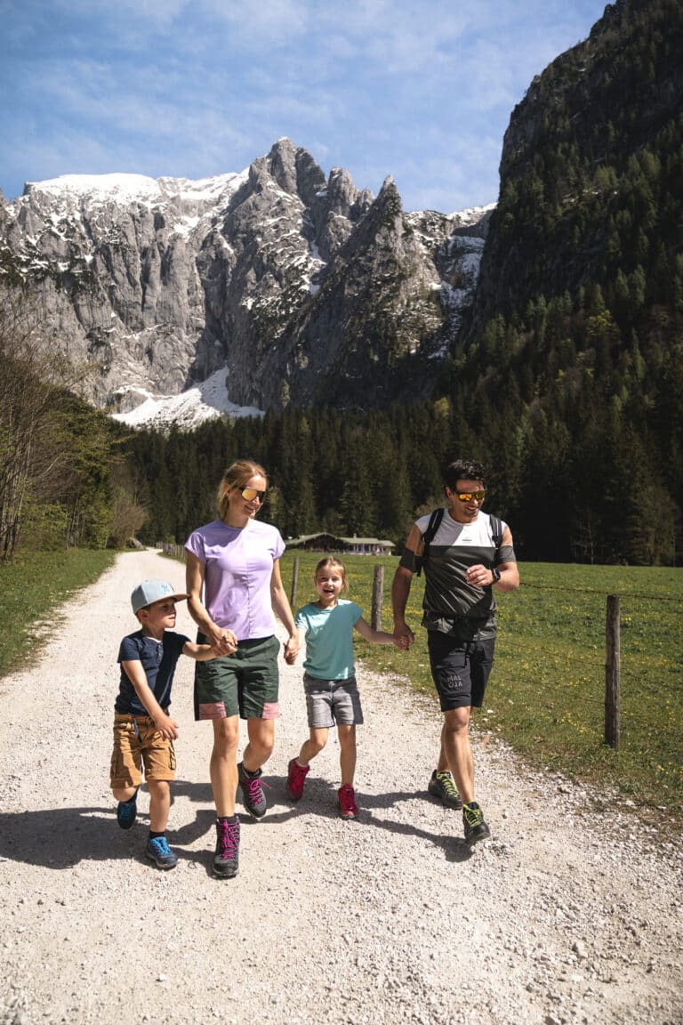 Eine vierköpfige Familie (Vater, Mutter und zwei Kinder) spaziert händchenhaltend fröhlich über einen Schotterweg durch ein sonniges Bergtal, vor grünen Wiesen, dichtem Nadelwald und schneebedeckten Felsgipfeln.