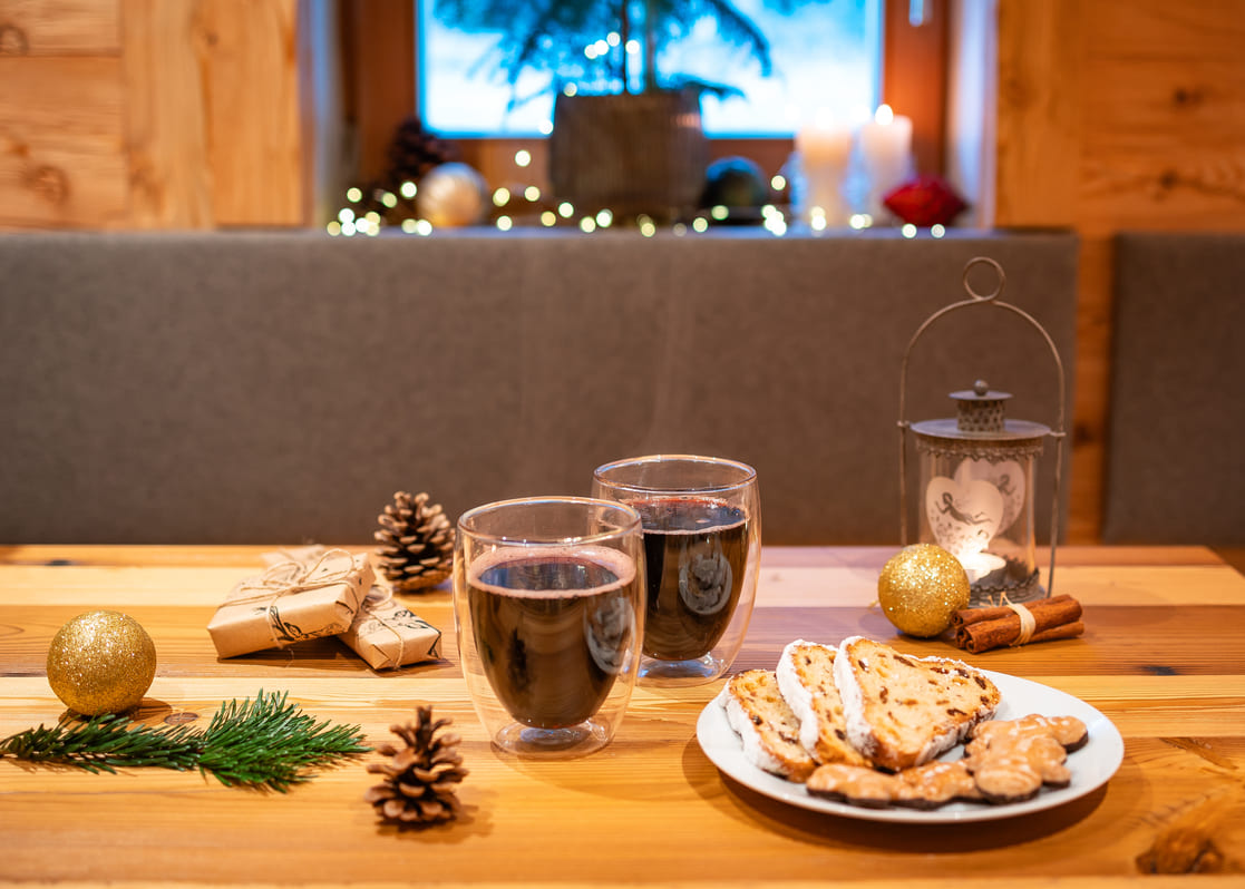 Zwei Gläser Glühwein auf einem Holztisch neben einem Teller mit Stollenscheiben und Weihnachtsplätzchen, umgeben von Tannenzweigen, Tannenzapfen, goldenen Kugeln, verpackten Geschenken, Zimtstangen und einer Laterne vor einem Fenster mit verschwommenen Lichterketten.