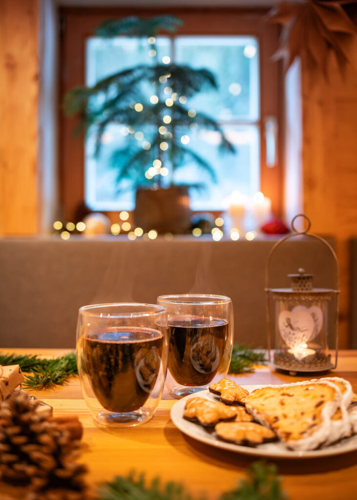 Zwei dampfende Doppelwandgläser mit dunklem Glühwein auf einem hölzernen Tisch, daneben ein Teller mit Weihnachtsgebäck und Stollen, Tannenzweige und Zapfen sowie eine brennende Laterne vor einem unscharf beleuchteten Weihnachtsbaum im Fenster.