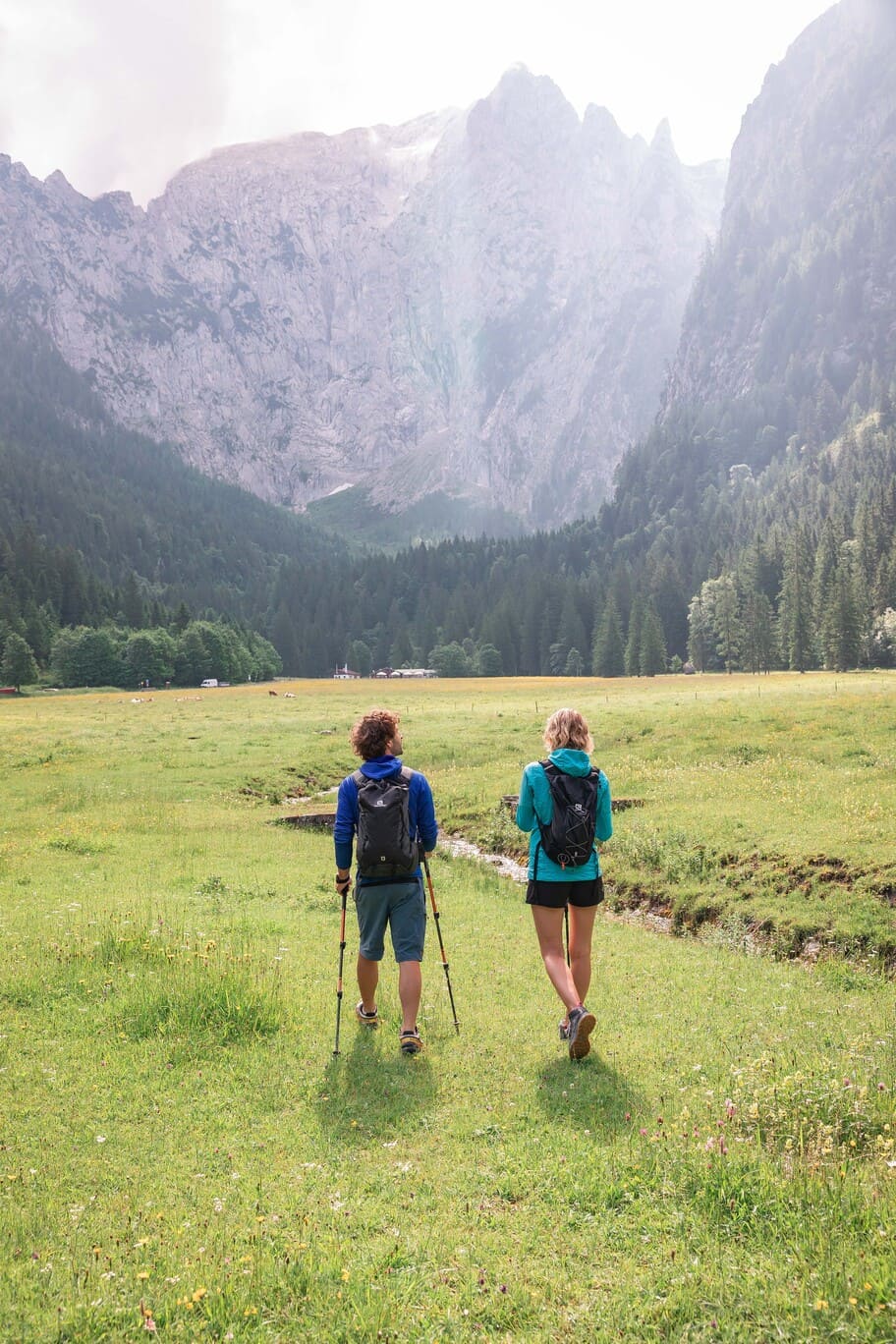 Zwei Wanderer mit Rucksäcken — ein Mann mit Trekkingstöcken und eine Frau in türkisfarbener Jacke — laufen über eine blühende Almwiese auf einen dichten Nadelwald und hohe, steile Felswände in einem Alpenkessel zu.