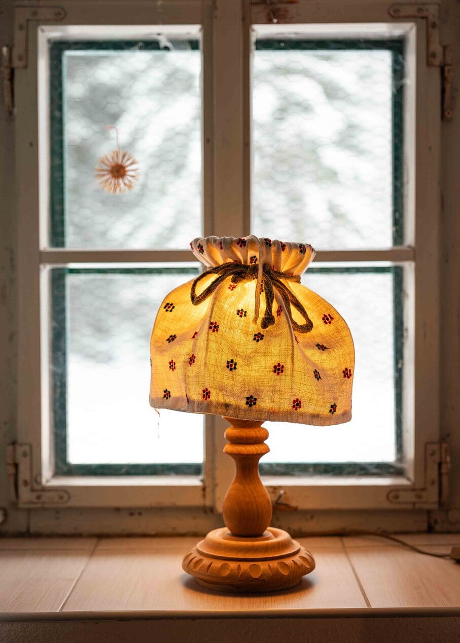 Tischlampe mit geschnitztem Holzfuß und gerafftem, besticktem Stoffschirm, die warm leuchtet und auf einer Fensterbank vor einem verschneiten, unscharfen Fenster steht.
