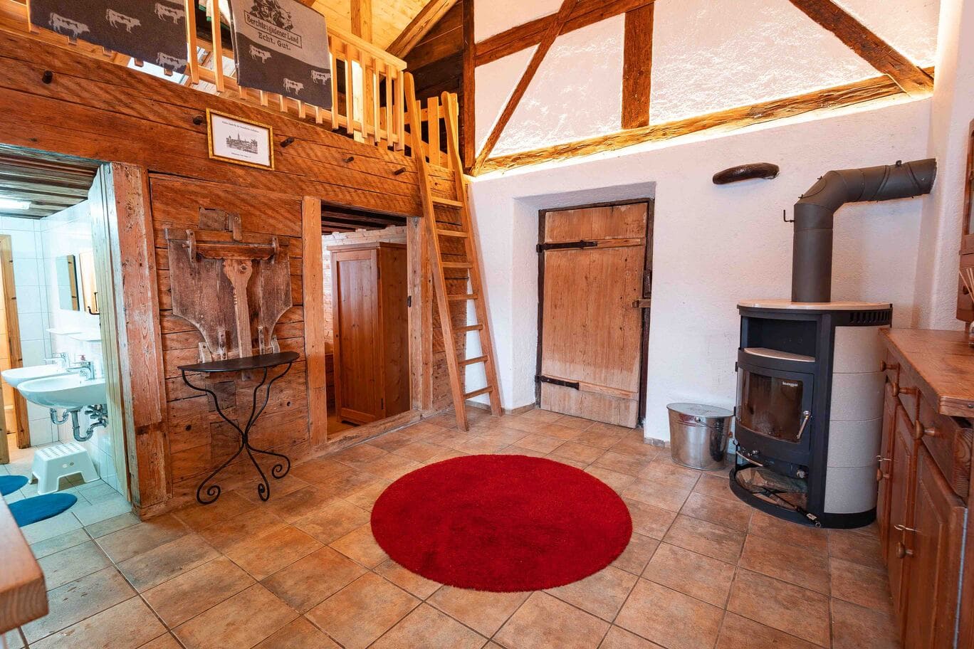 Innenraum der Scharitzkehlalm: rustikale Diele mit sichtbaren Holzbalken, einer Holzleiter zur Empore, rundem roten Teppich, gusseisernem Holzofen, Holztür und Blick in ein kleines Bad mit Waschbecken.