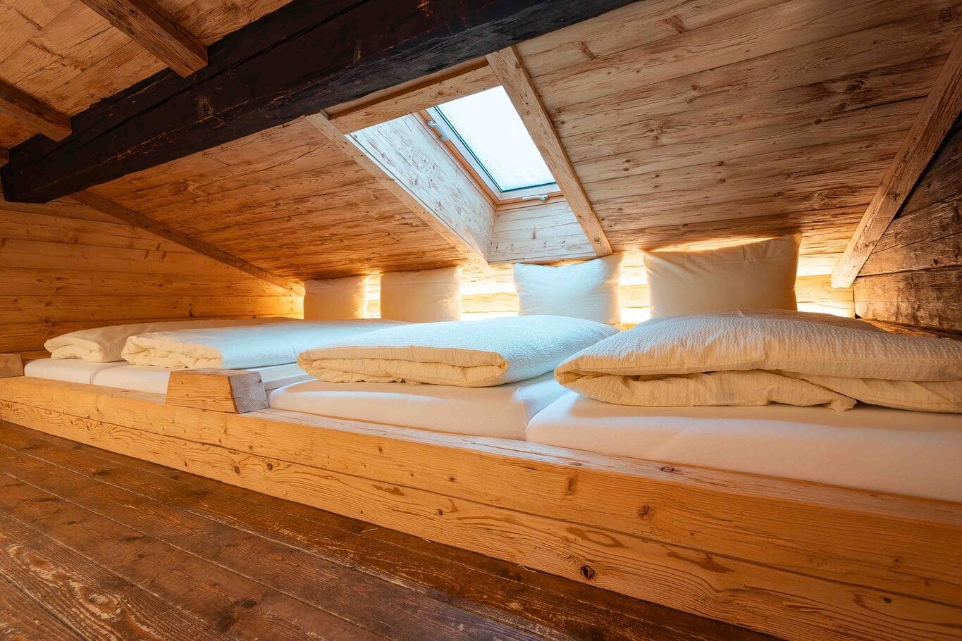 Gemütliche Schlafgalerie in einer Almhütte mit niedriger, schräger Holzbalkendecke, drei nebeneinander liegenden Matratzen mit weißen Decken und Kissen, warmem indirektem Licht und einem Dachfenster.