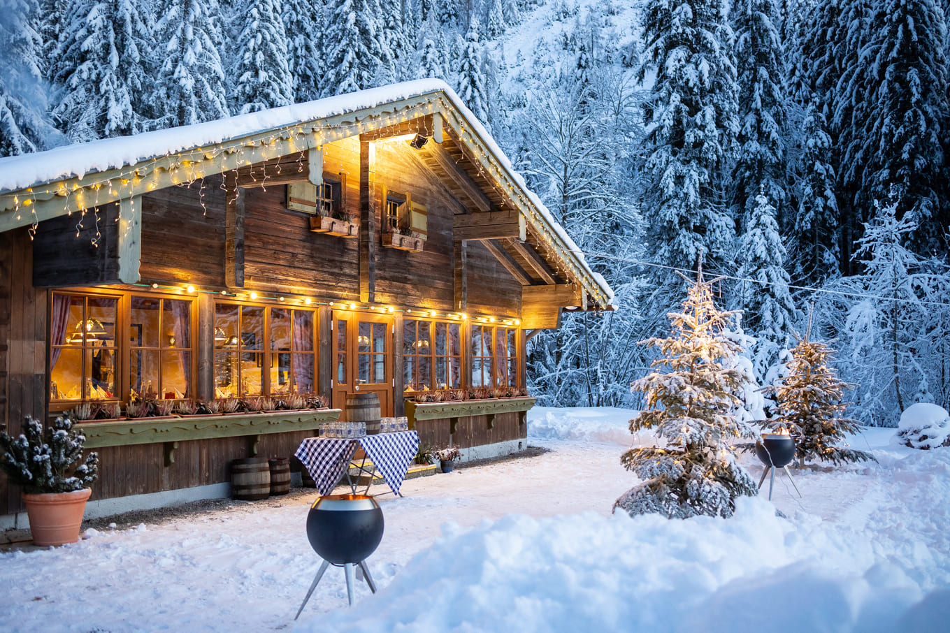 Beleuchtete Holzhütte im verschneiten Tannenwald bei Dämmerung, mit Lichterketten am Dach, warmem Innenlicht durch große Fenster, geschmückten schneebedeckten Tannen und zwei runden Feuerschalen auf dem verschneiten Vorplatz.